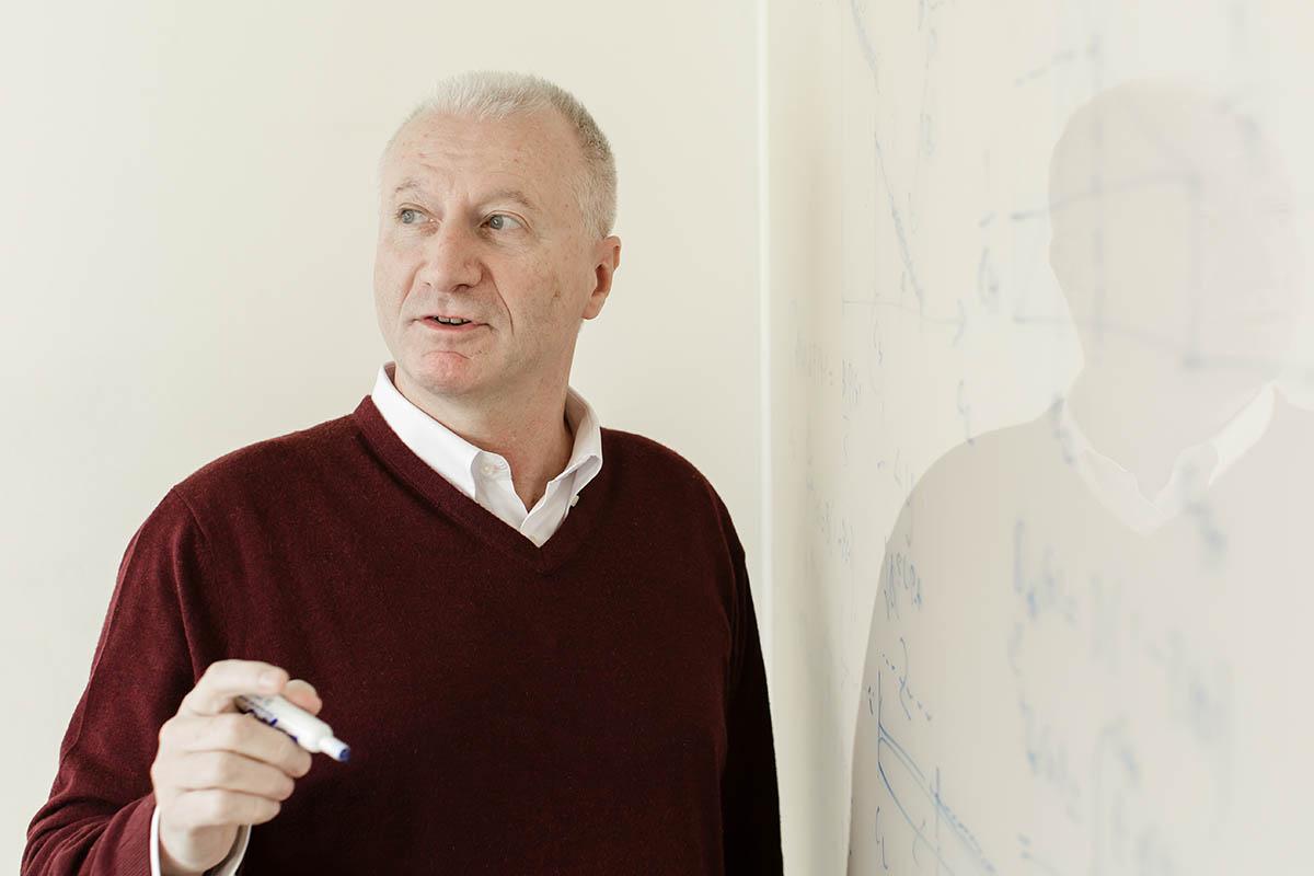 Professor Giuseppe "Pino" Lopomo at Duke University's Fuqua School of Business, he is wearing a white colored shirt with deep red sweater, standing to the left of a white boarding holding a blue dry-erase marker and looking to his right